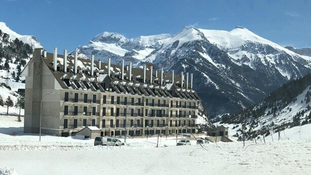 Un hotel de varios pisos con un diseño arquitectónico moderno, que cuenta con una chimenea central destacada y una fachada simétrica. El hotel está ubicado en una zona montañosa nevada con cielos azules despejados y picos nevados al fondo.