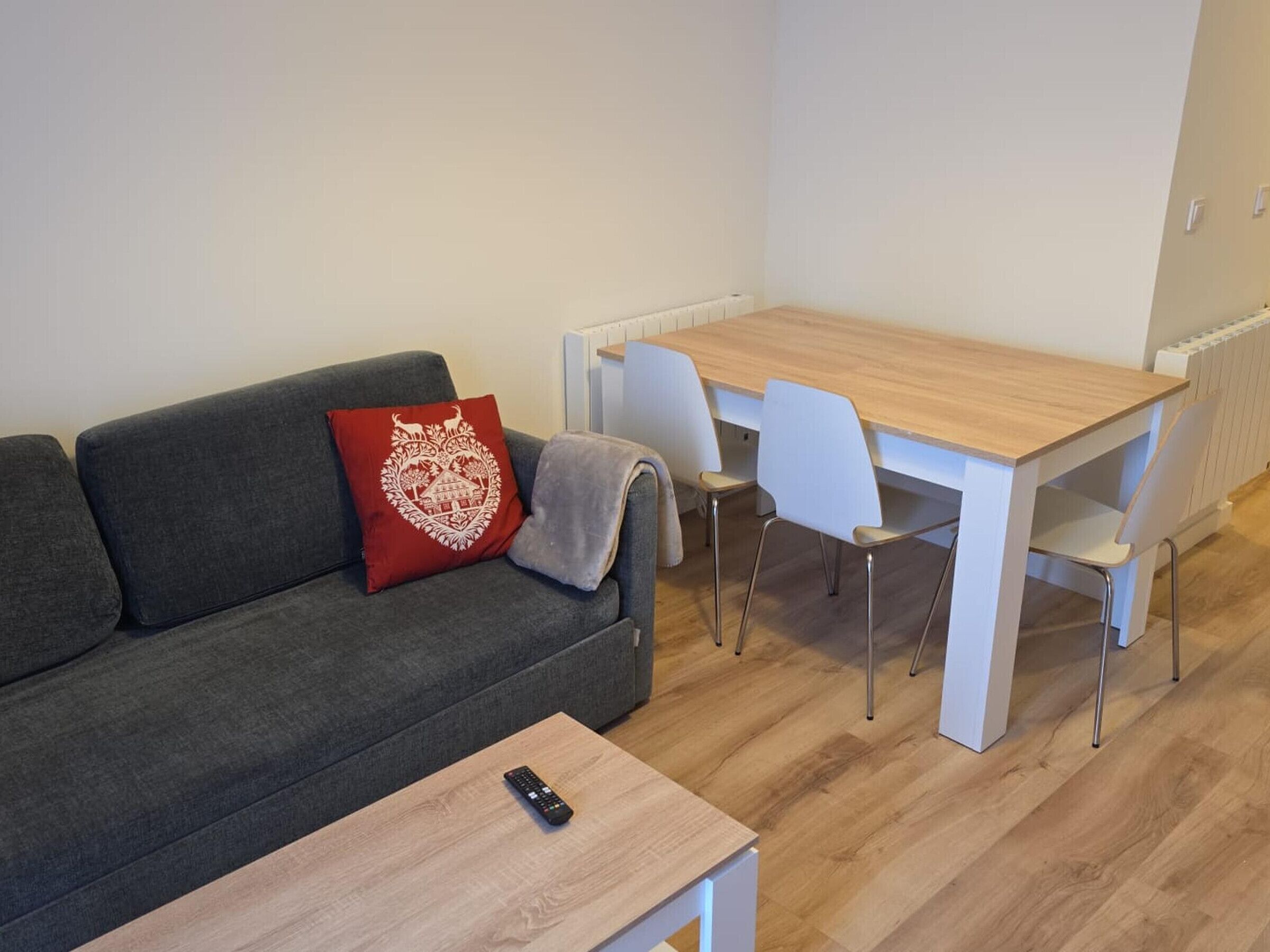 A modern living room with a grey sofa, a red decorative pillow, a wooden coffee table, and a white dining table with chairs.