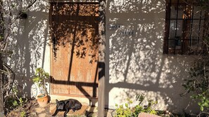 Eine rustikale Holztür, vor der ein Hund liegt, umgeben von Grünpflanzen und einer Mauer mit der Aufschrift "MOZA LUZ".