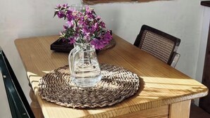 A wooden dining table with a woven placemat, a vase with purple flowers, and a window with wooden shutters.