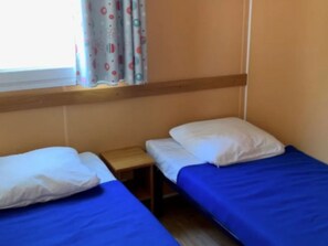 A small, single-room accommodation with two single beds, a wooden nightstand, and a window with patterned curtains. - Mobile Home with Terrace for 4 People (Arvert)