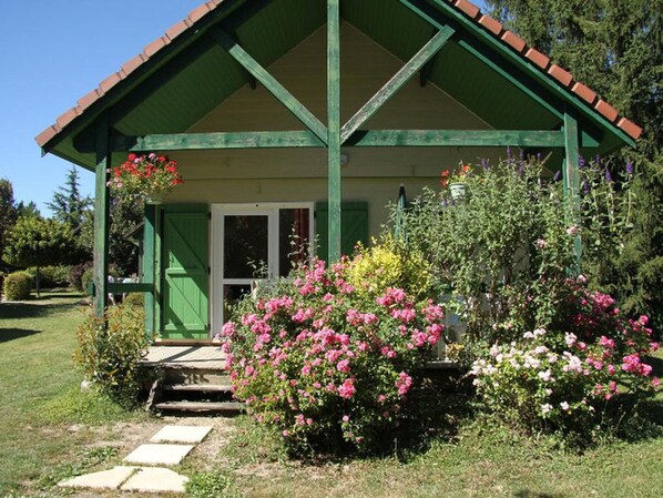 A small, green-roofed building with a porch, surrounded by lush gardens and flowering plants.