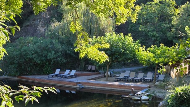 Un área de piscina tranquila al aire libre con sillones de descanso y una terraza de madera sobre el agua, rodeada de abundante vegetación.