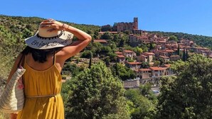 Una persona con un vestido amarillo y sombrero blanco observa un pueblo en una ladera con edificios históricos y una torre destacada.