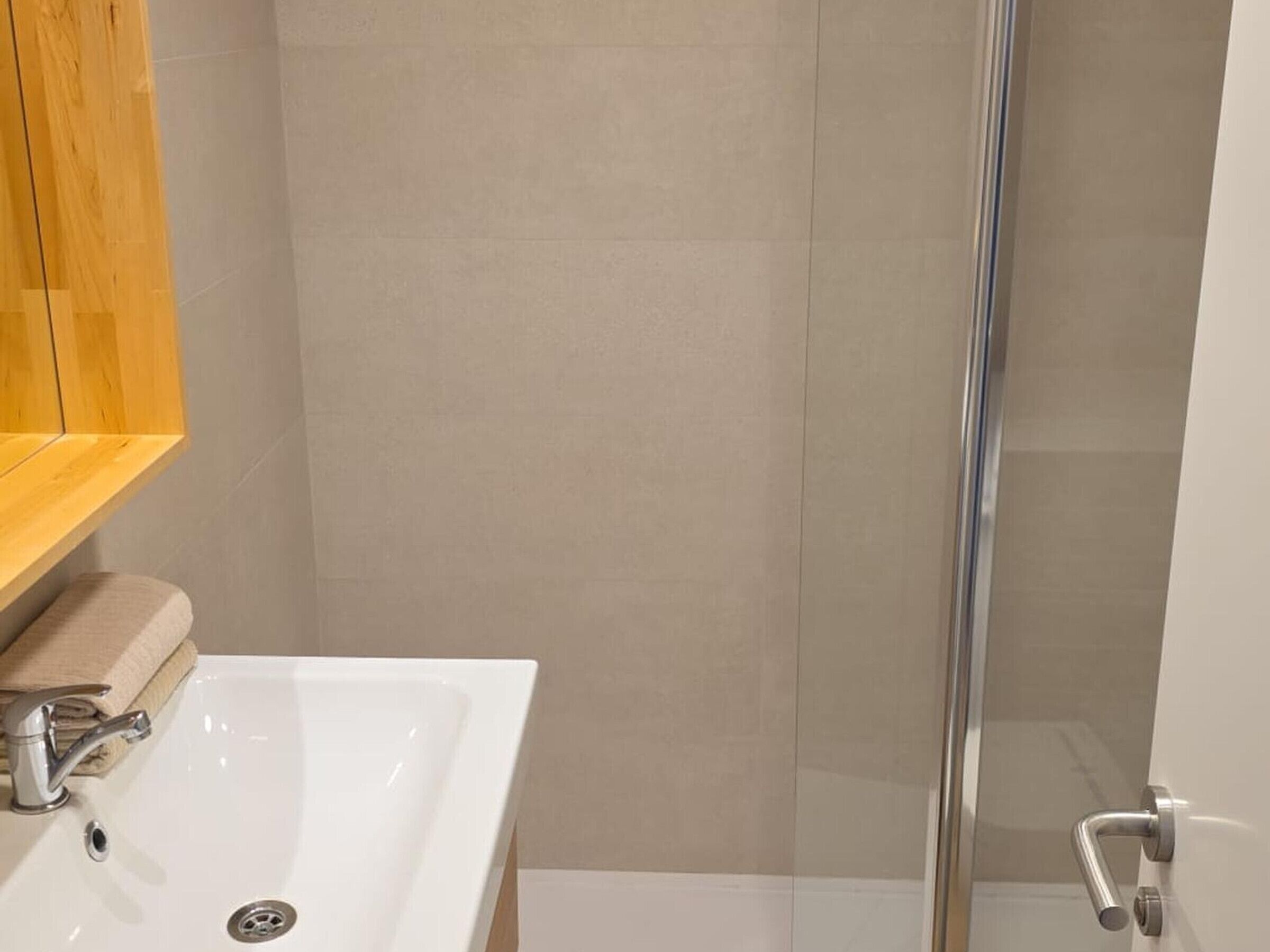 A bathroom with a white sink, a wooden shelf, and a glass shower enclosure.