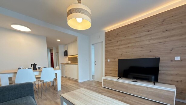 Een moderne woonkamer met een flatscreen televisie, een houten accentmuur, en een eethoek met witte stoelen, met een keuken op de achtergrond.