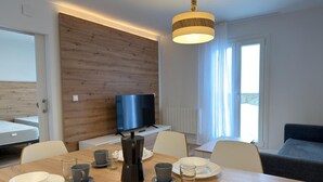 Een moderne eethoek met een houten tafel, stoelen en een koffiezetapparaat. Een televisie is aan de muur bevestigd, en er is een raam met gordijnen.