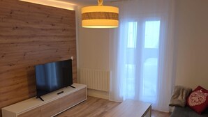 Een moderne woonkamer met een flatscreen televisie op een houten kast, een klein tafeltje, een bank met rode kussens en een groot raam met dunne gordijnen.
