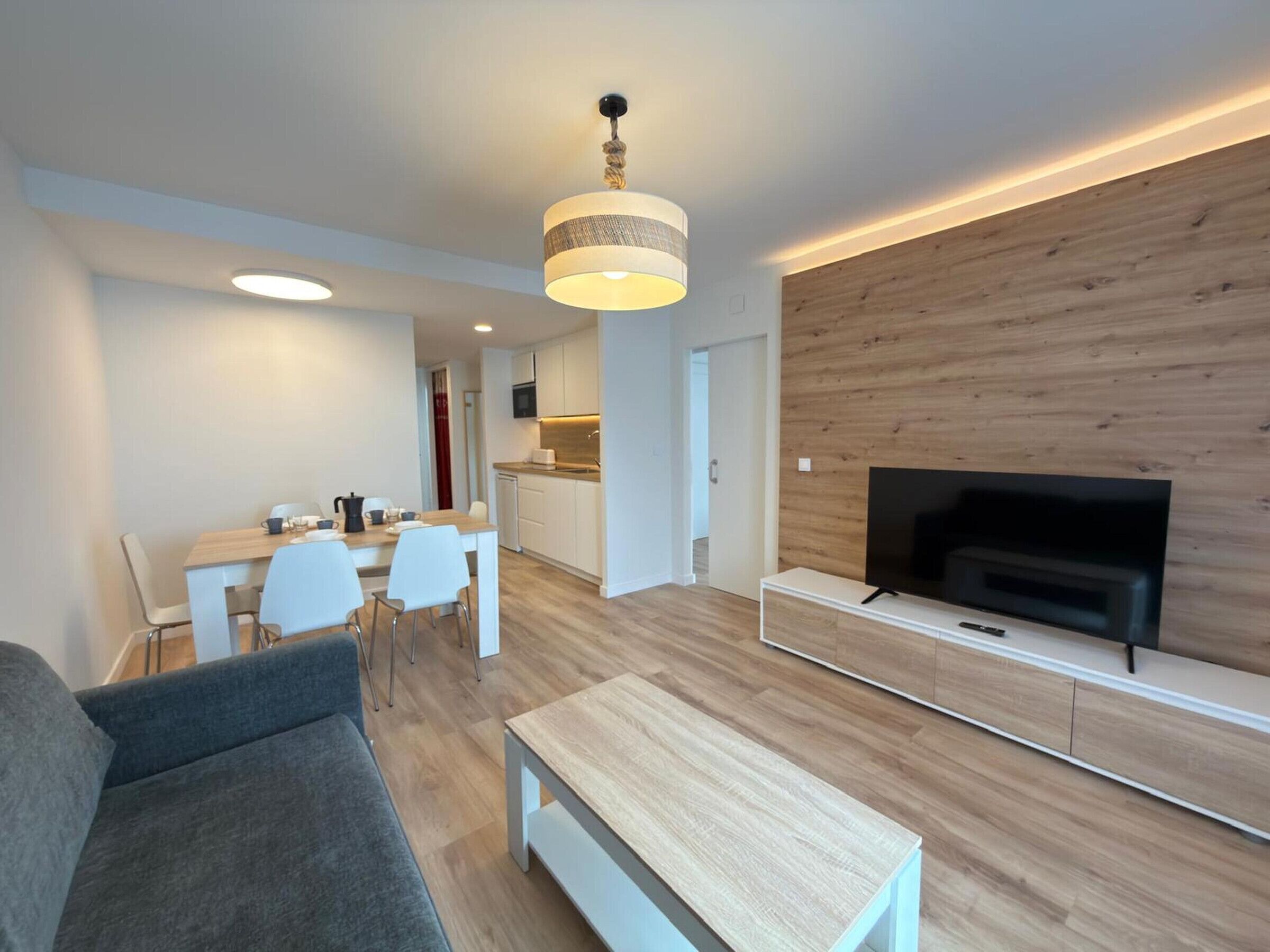 A modern living room with a sofa, a coffee table, a TV on a wooden wall, a dining area with chairs and a table, and a kitchen with white cabinets and a microwave.