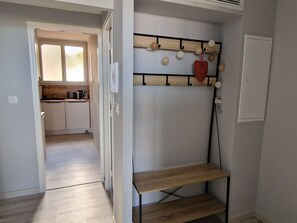 A narrow hallway with a wooden shelf unit and a wall-mounted light fixture. A kitchen is visible in the background with a window and cabinets. - Quiet T2 Apartment, 4 Pers, Parking (Banyuls-sur-Mer)