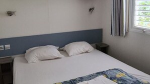 A single bed with a blue headboard, white pillows, and a floral-patterned blanket. There is a small bedside table with a lamp, a window with curtains, and a wall-mounted shelf above the bed. - Mobile home 27sqm, 6 pers, 3 bed, terrace (Espira-de-Conflent)