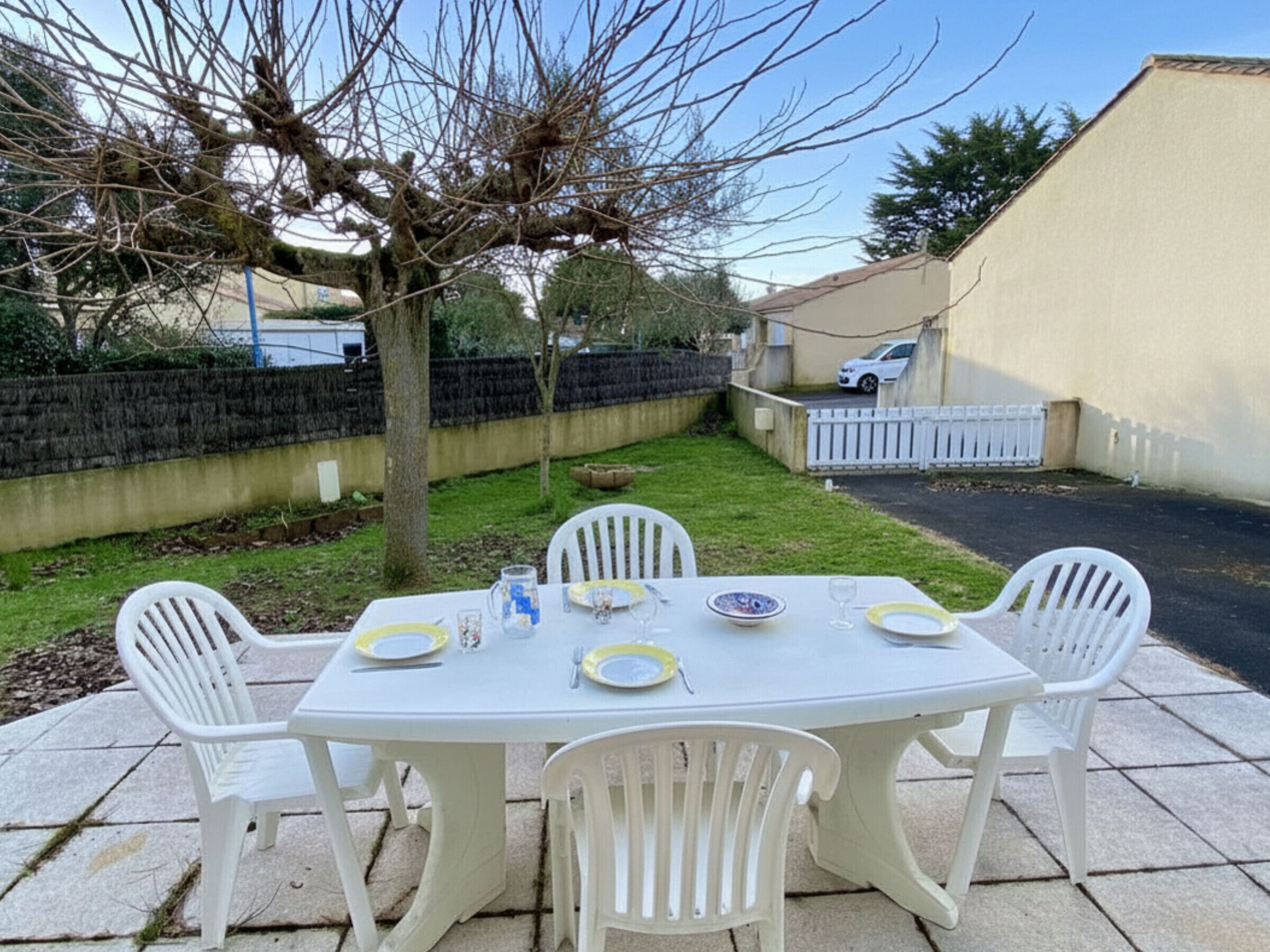Une terrasse avec une table dressée pour quatre personnes, entourée de chaises en plastique blanches, d’un arbre et d’une clôture blanche.