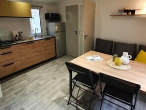 A compact kitchen with wooden cabinets, a microwave, and a refrigerator. There is a dining table with chairs and a small shelf above it. - Mobile Home with Terrace for 4 People (Arvert)