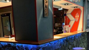 Eine Barbereitschaft mit einer Steintheke und Hockern, einem Kühlschrank und einer Wand mit geometrischem Muster sowie einem Poster.