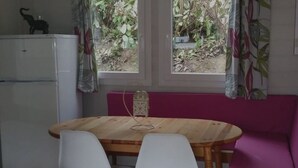 Eine kleine Küche mit Holztisch, zwei weißen Stühlen, einem Kühlschrank und einem Fenster mit Vorhängen.