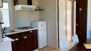 A compact kitchen with a gas stove, a refrigerator, and a dining area with a table and chairs. - Cosy mobile home with terrace for 4 (Arvert)