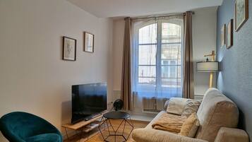 Un salon avec un téléviseur à écran plat posé sur un meuble en bois, un fauteuil turquoise, un canapé beige et une fenêtre avec des rideaux.