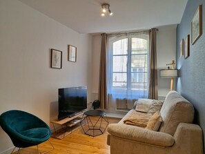 A living room with a flat-screen TV on a wooden stand, a teal chair, a beige sofa, and a window with curtains.