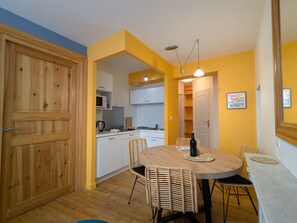 Cocina compacta con gabinetes blancos, microondas y estufa. Hay un comedor con una mesa redonda y sillas de mimbre.