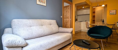 Una sala de estar con un sofá, una mesa de centro redonda y una silla. Se ve un área de cocina al fondo.