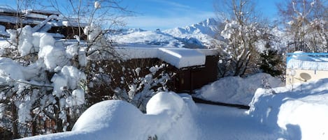 A snow-covered landscape with a traditional wooden building, a mountain range in the background, and a clear blue sky.