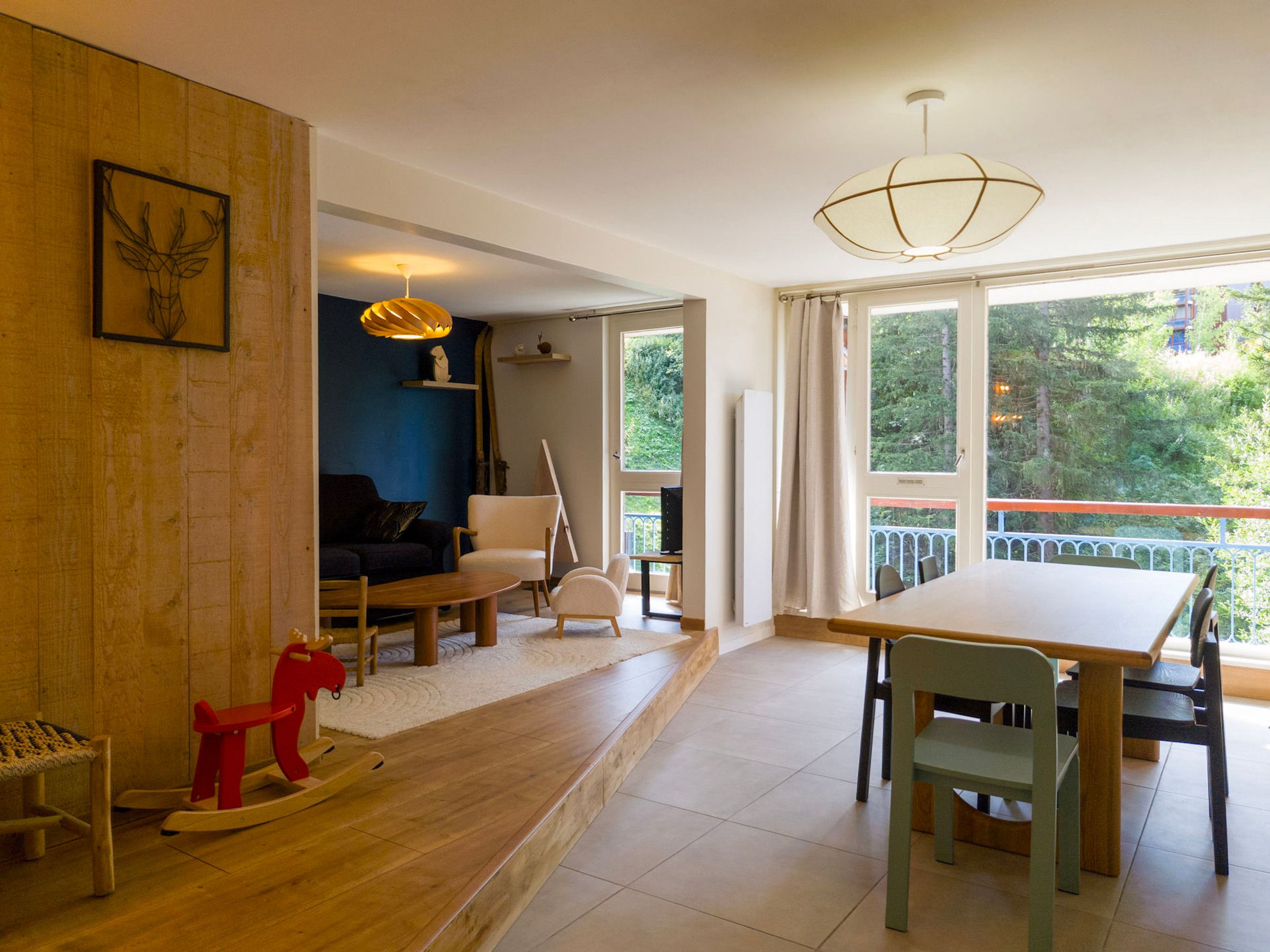 A modern living room with a dining area, a wooden floor, and a view of greenery outside.