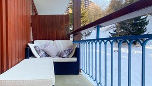 En balkong med en vit soffa och kuddar, ett blått räcke och utsikt över ett snötäckt landskap.