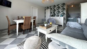 Een moderne woonkamer met een eethoek, een grijze bank, rieten stoelen, een witte salontafel en een keuken op de achtergrond.