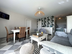 A modern living room with a dining area, a grey sofa, wicker chairs, a white coffee table, and a kitchen in the background. - 3-Room Apartment with Balcony and Pool in Coëx (Coëx)