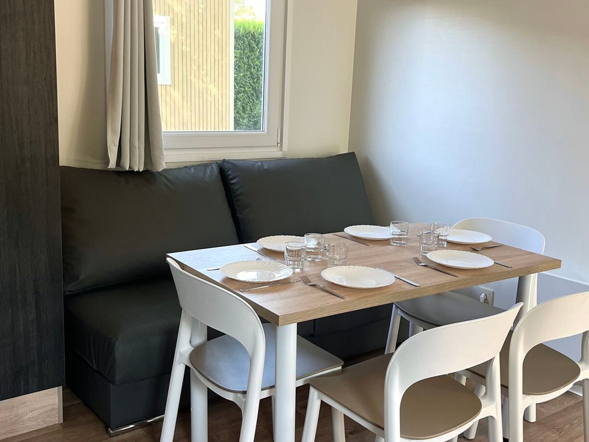 Un comedor con una mesa de madera preparada para cuatro personas, sillas blancas, un sofá gris oscuro y una ventana con cortinas.