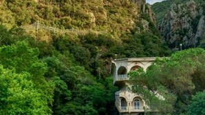 Un edificio de piedra con ventanas de arco, rodeado de abundante vegetación y terreno rocoso.
