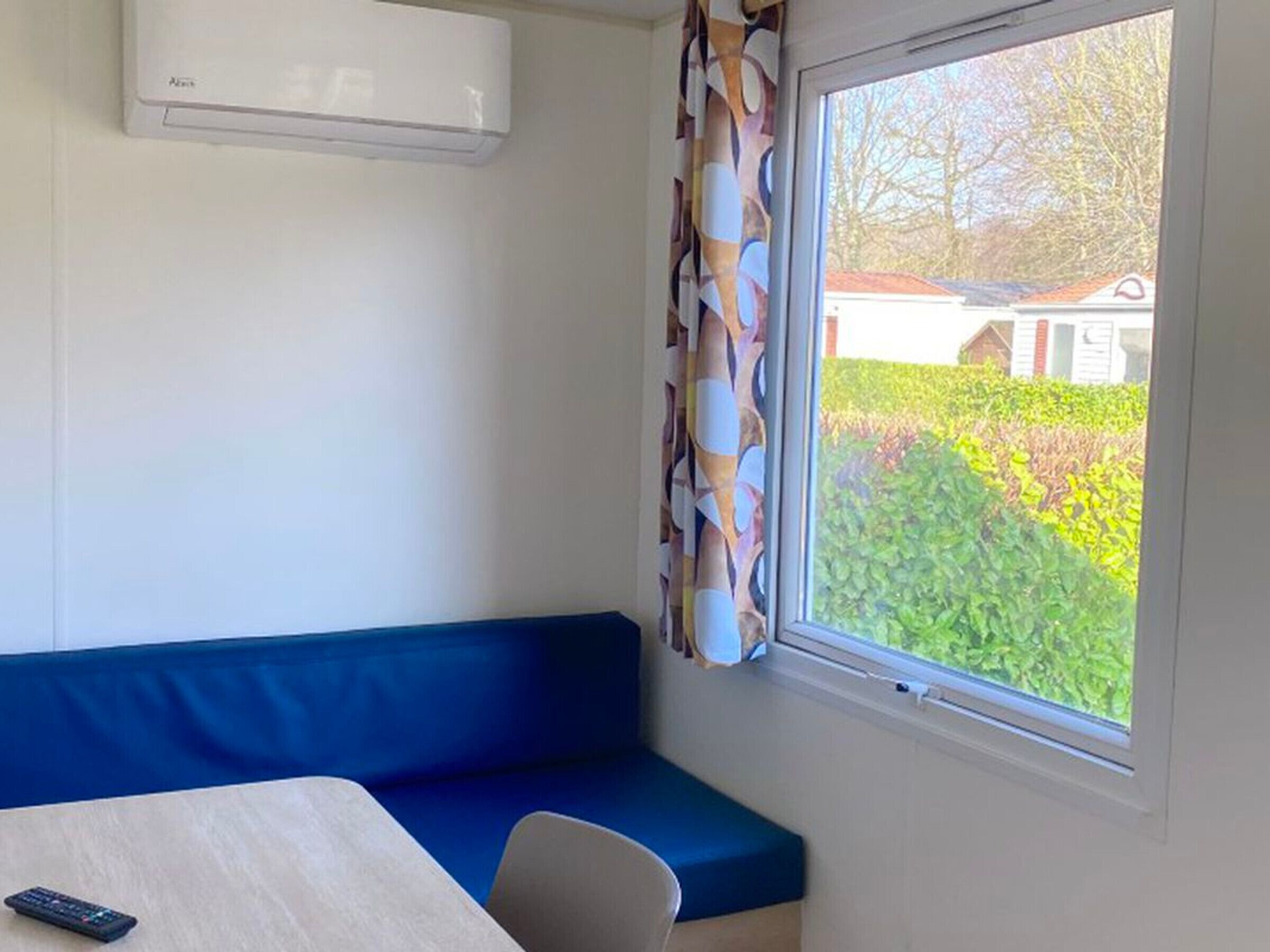 A room with a blue sofa, a table, a remote control, and a window with a view of greenery and a building.