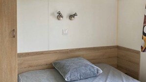 A bedroom with a wooden headboard, a single bed with a grey pillow, and a white wardrobe with two metallic handles. - 20m² Mobile Home for 2 Guests (Angicourt)