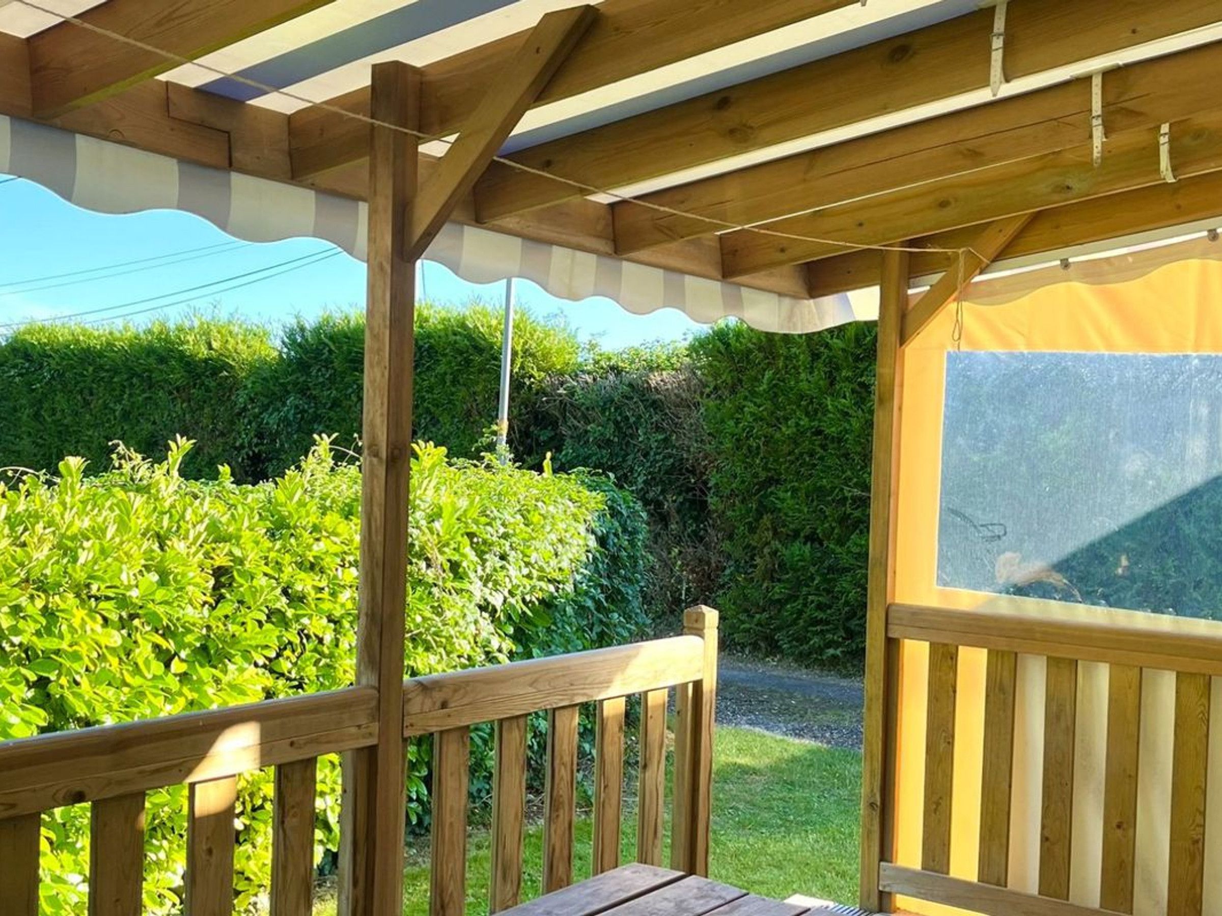 Une terrasse en bois avec un banc, un espace abrité doté d’un auvent blanc, et une vue sur une haie et une route.