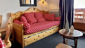 A cozy room with a wooden sofa bed covered in red and white heart-patterned bedding, a small round table with a white vase, a mirror on the wall, and a window with curtains. - Apartment at slopes, Wifi included, sauna, 5 sleeps (Risoul)