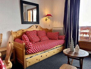 A cozy room with a wooden sofa bed covered in red and white heart-patterned bedding, a small round table with a white vase, a mirror on the wall, and a window with curtains. - Apartment at slopes, Wifi included, sauna, 5 sleeps (Risoul)