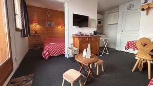 A cozy room with a bed, a dining table with chairs, a small wooden stool, a TV, and a kitchen area with a microwave. - Apartment at slopes, Wifi included, sauna, 5 sleeps (Risoul)