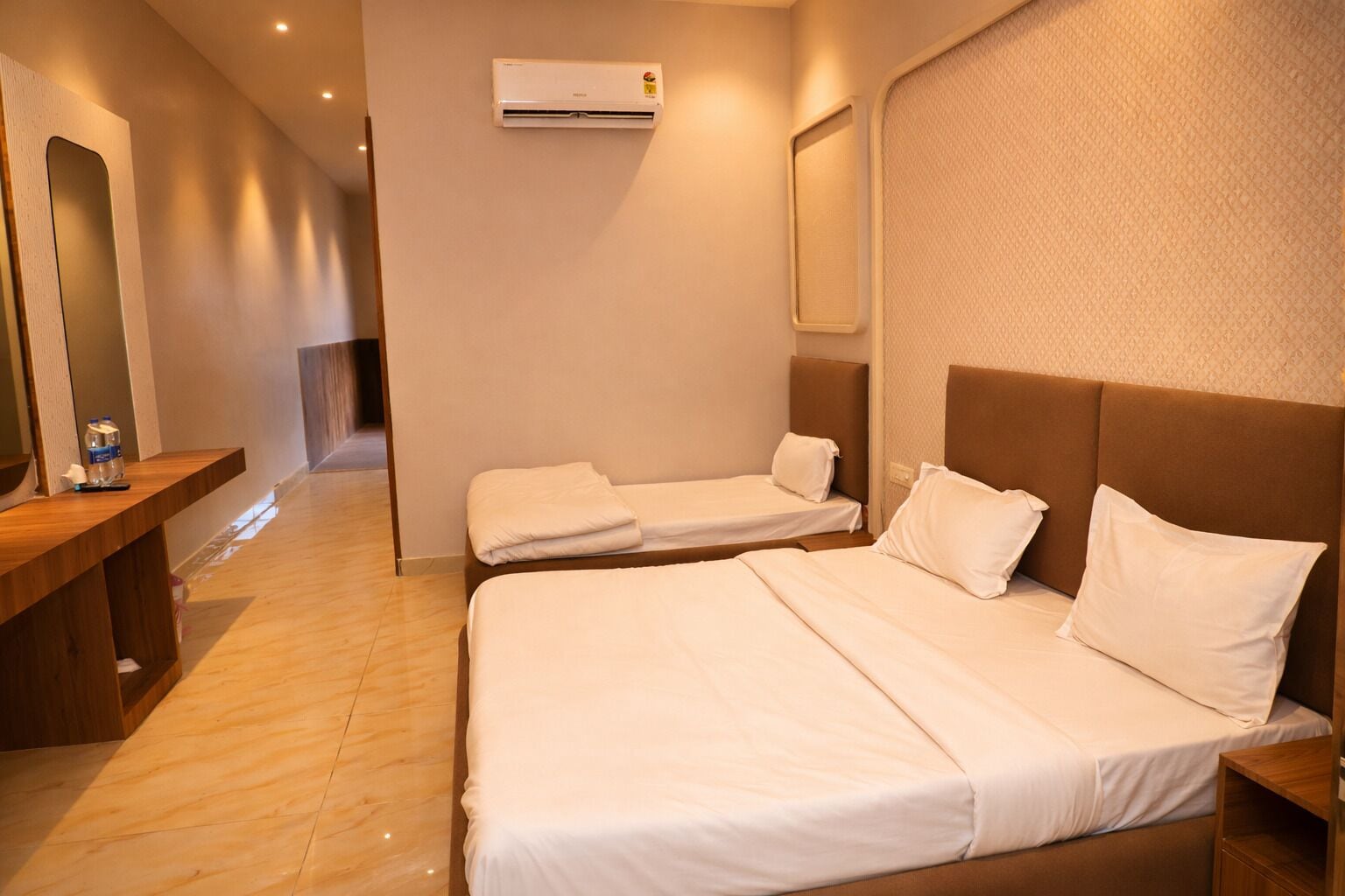 Una habitación de hotel con una cama grande, dos camas más pequeñas, un banco de madera, un espejo y un equipo de aire acondicionado.
