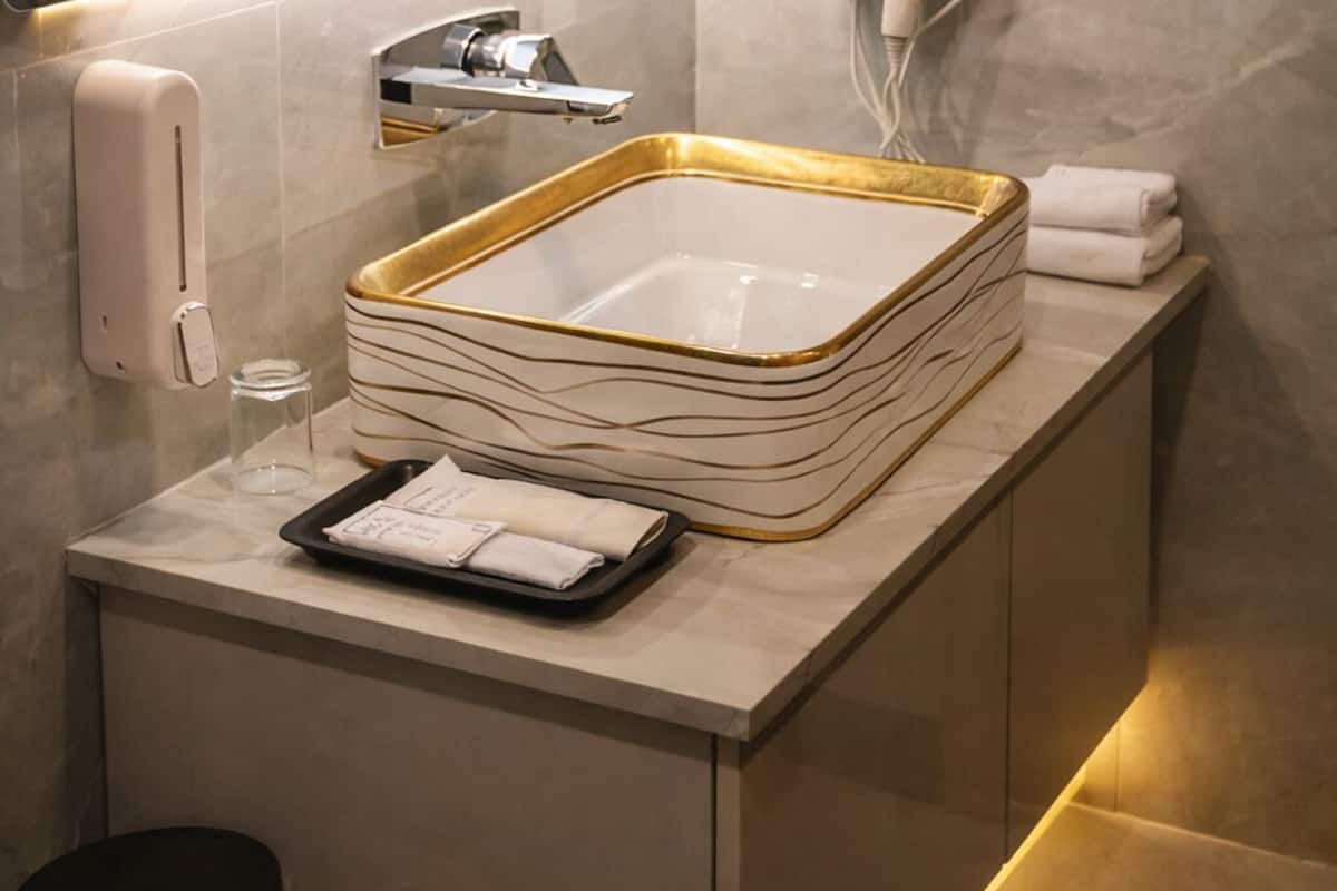 Une salle de bain moderne avec un grand miroir, un lavabo encadré d’or, un sèche-cheveux et un distributeur de savon.