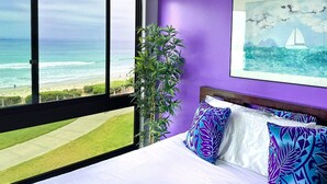 A bedroom with a large window overlooking a beach, a bed with patterned pillows, and a painting of a sailboat. - Ocean Views * Corner Unit * POOL & HOT TUB (San Diego)