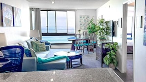 A modern living room with a sofa, coffee table, and chairs, a dining area with a table and chairs, and a large window with a view of the ocean. - Ocean Views * Corner Unit * POOL & HOT TUB (San Diego)