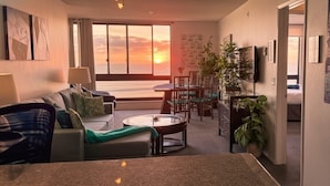 A living room with a sofa, coffee table, and dining area with chairs. There is a large window with a view of the sunset. - Ocean Views * Corner Unit * POOL & HOT TUB (San Diego)