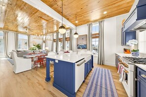A modern kitchen with a central island, wooden ceiling, and large windows. - Designer Coastal Cottage with Lake Views and 3 King Suites at Cinnamon Shore (Port Aransas)