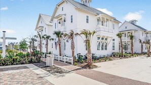A large white building with multiple windows and a balcony, surrounded by palm trees and a paved walkway. - Designer Coastal Cottage with Lake Views and 3 King Suites at Cinnamon Shore (Port Aransas)