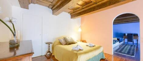 A bedroom with a wooden ceiling, a bed with a yellow bedspread, a nightstand, a vase with flowers, and a view through an archway to another room.