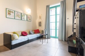 Ein Wohnzimmer mit einer Couch, einem kleinen Tisch, gerahmten Pflanzenbildern an der Wand und einem Fenster mit Vorhängen.