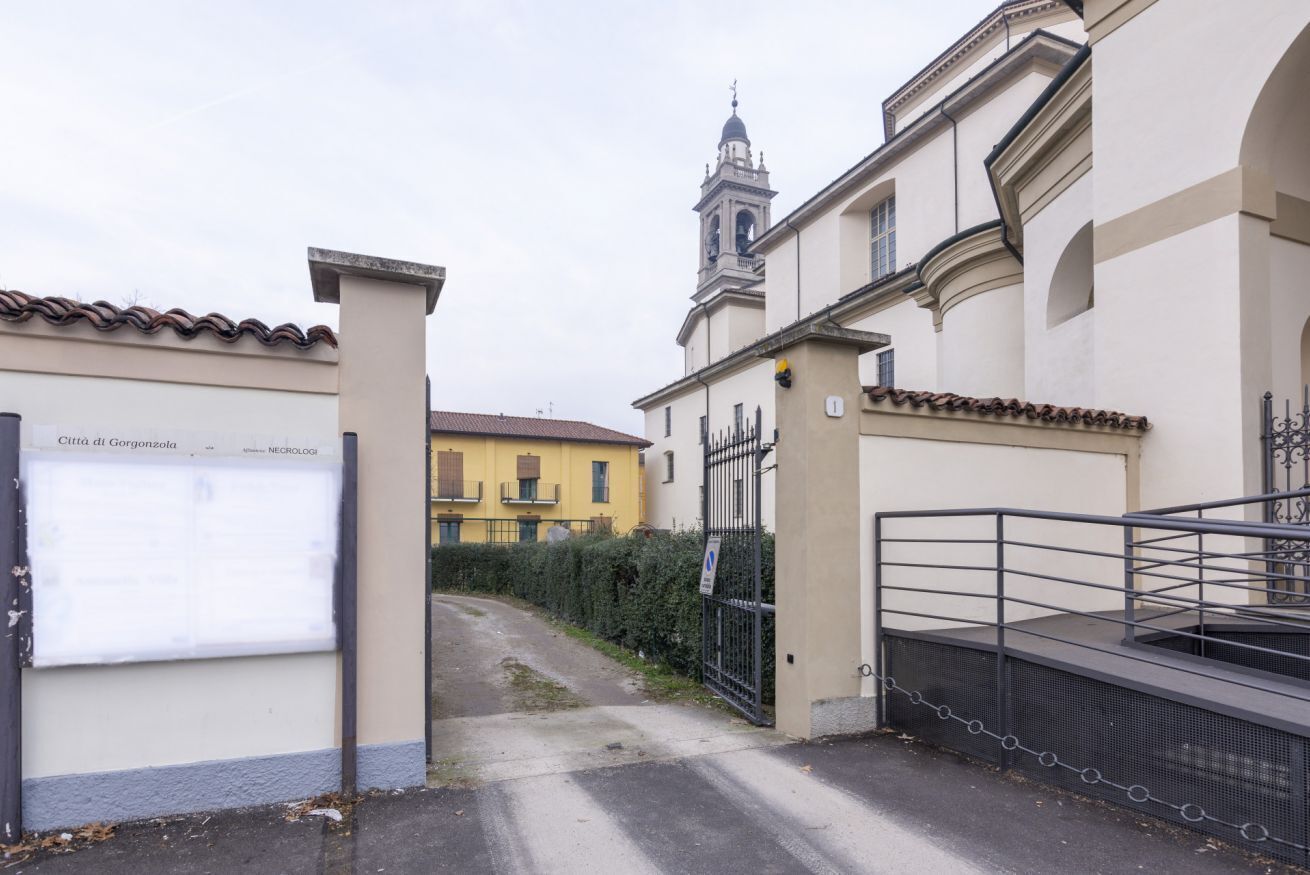 Une entrée fermée par un portail mène à un bâtiment doté d'un clocher, un panneau indique « Ville de Gorgonzola », et un chemin pavé conduit à l'entrée.