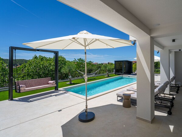 A modern outdoor pool area with a swing, lounge chairs, and a large umbrella.