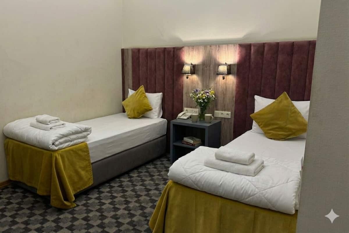 Habitación de hotel con dos camas individuales, cada una con una colcha amarilla y almohadas blancas. Hay una mesita con un teléfono y un jarrón de flores. El suelo es de cuadros y el cabecero de madera.