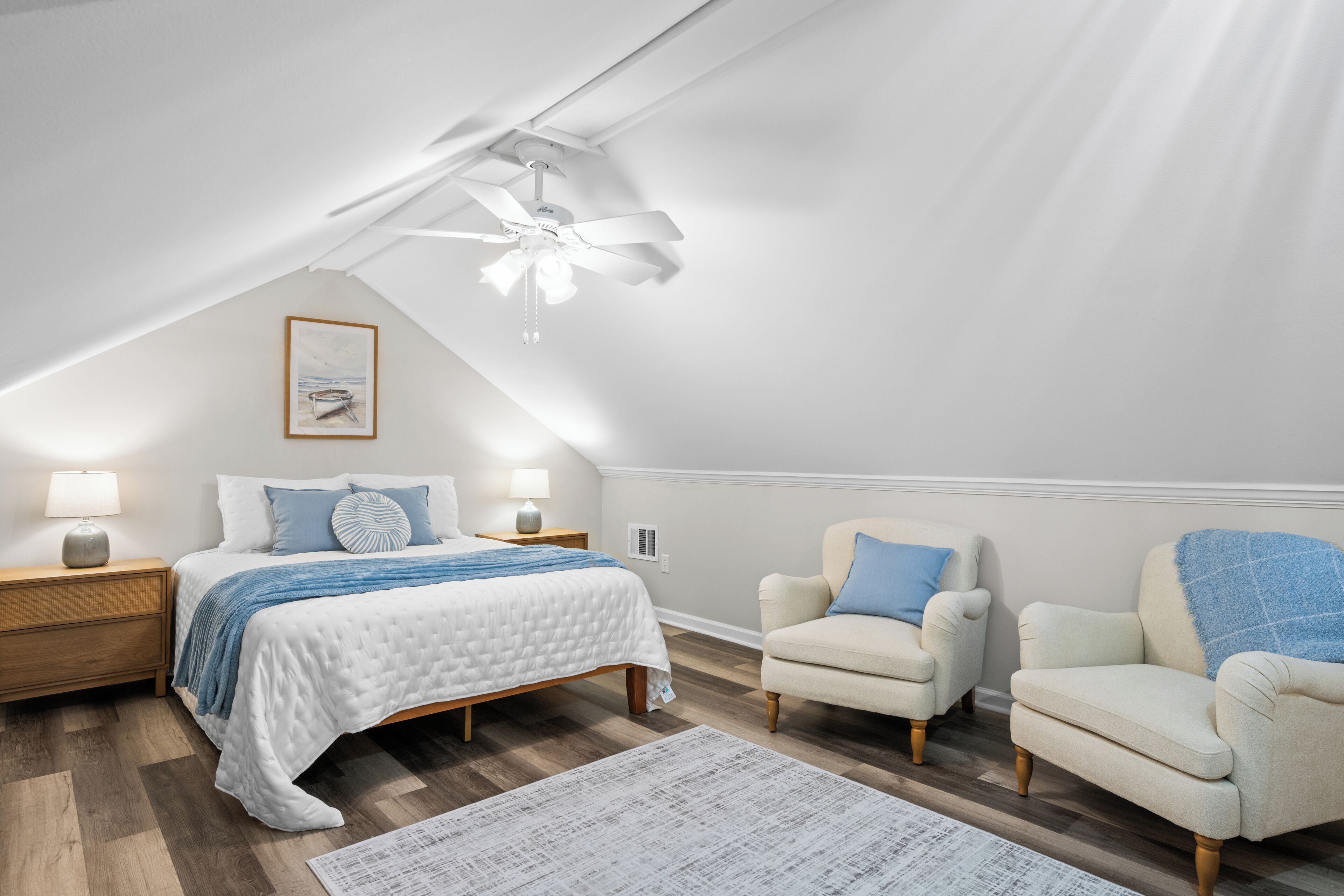 A bedroom with a bed, two chairs, a nightstand, a ceiling fan, and a framed picture on the wall.